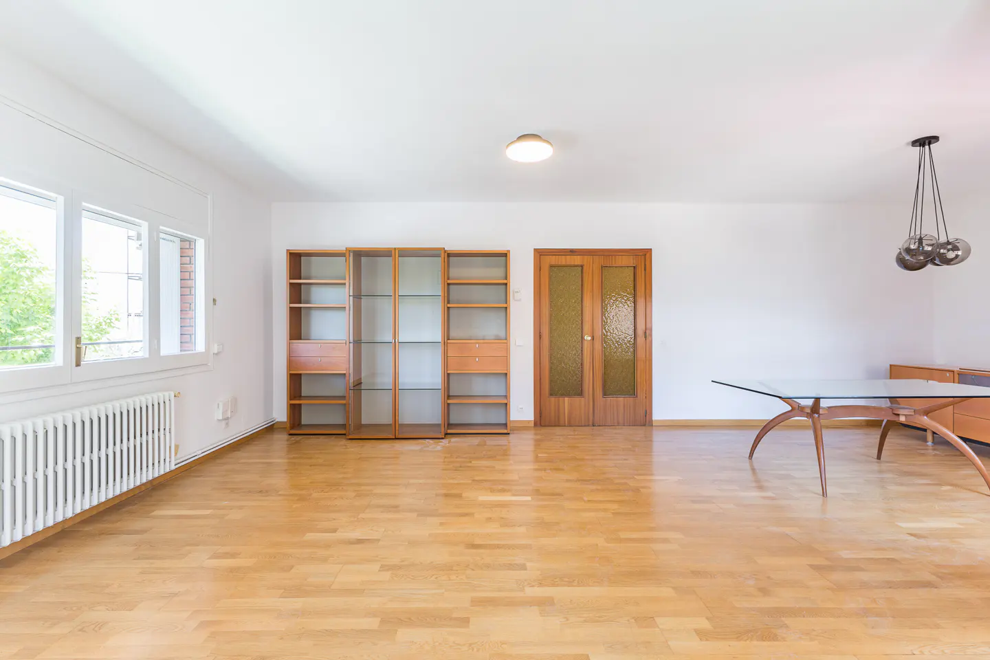 Bright, empty living room with hardwood floors, white walls, and built-in wooden shelving. A glass table and modern light fixture are visible.