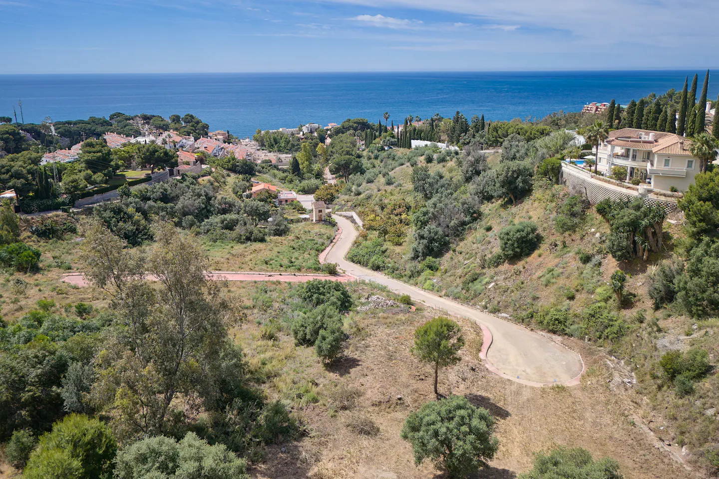 Hilly landscape with a winding road leading to a luxury villa overlooking the blue ocean and sky. Green trees and shrubs surround the property.