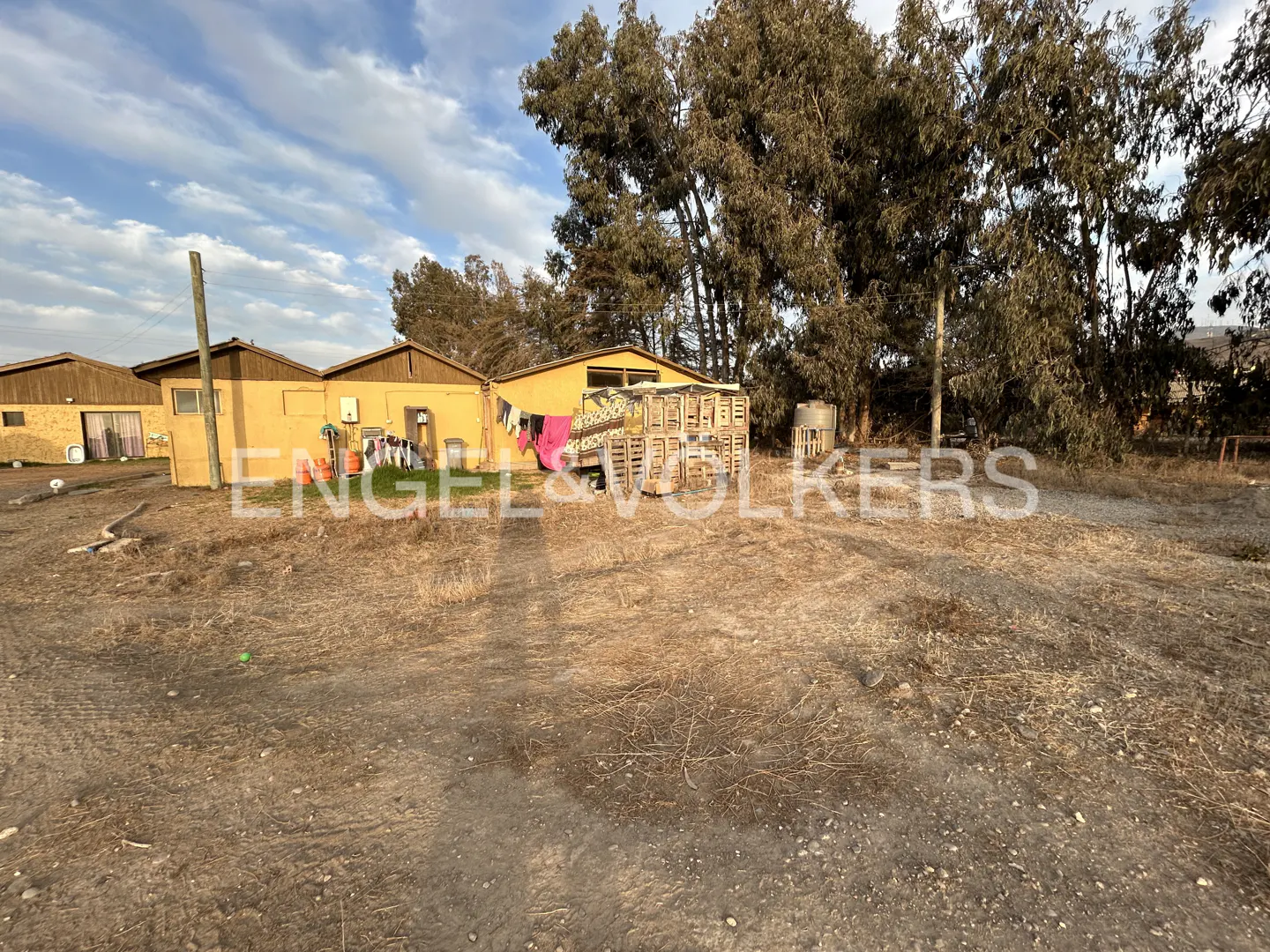 Yellow rural house with laundry hanging outside, wooden crates, and tall trees under a blue sky with light clouds.