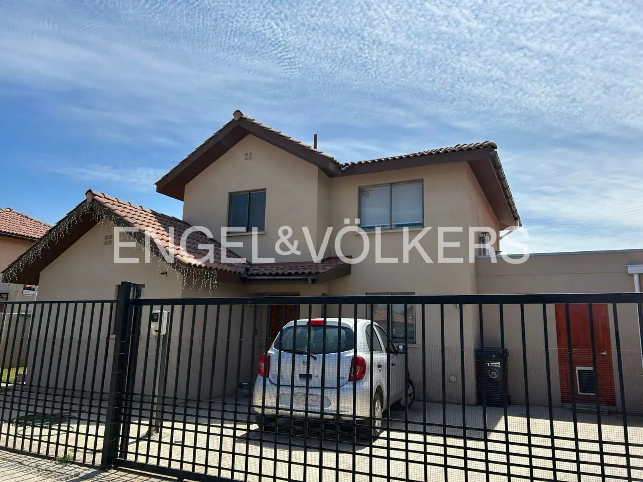 Two-story beige house with a red tile roof and black iron fence. A silver car is parked in the driveway. Engel & Völkers logo on the house.