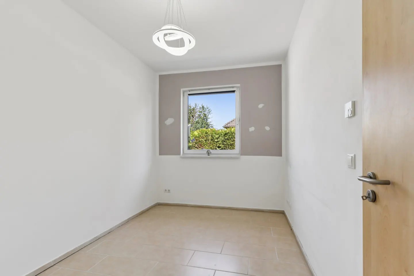 Bright, empty room with white walls, beige tile floor, and a window showing green foliage. A modern light fixture hangs from the ceiling.