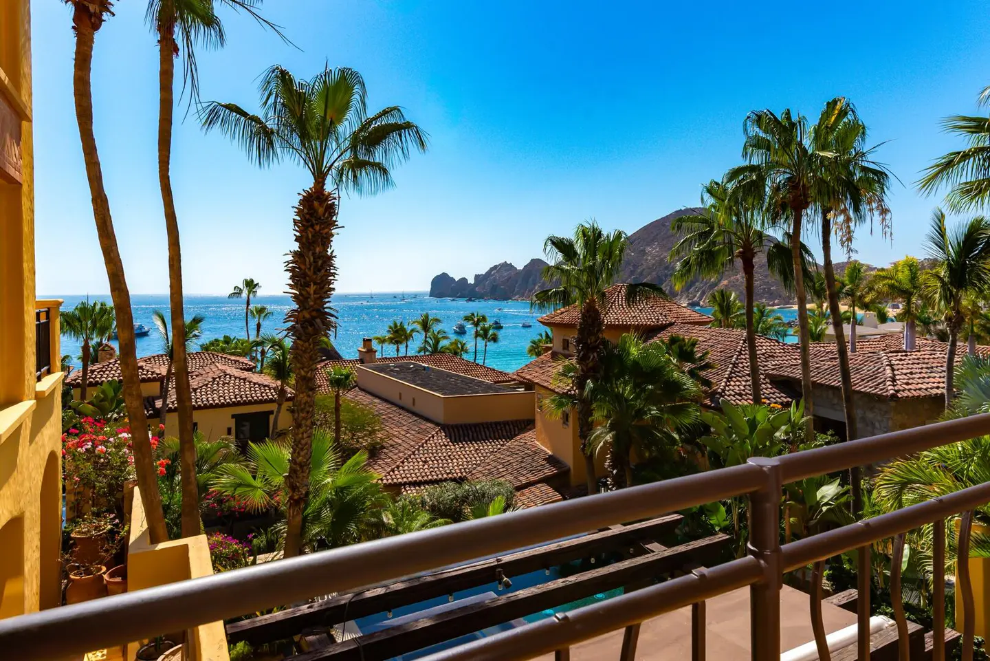 View from a balcony overlooking tropical villas with red tile roofs, palm trees, and the ocean under a clear blue sky.