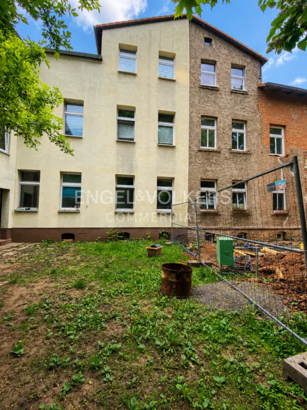 Exterior view of a multi-story building with cream and stone facades, a grassy yard, and a temporary construction fence.