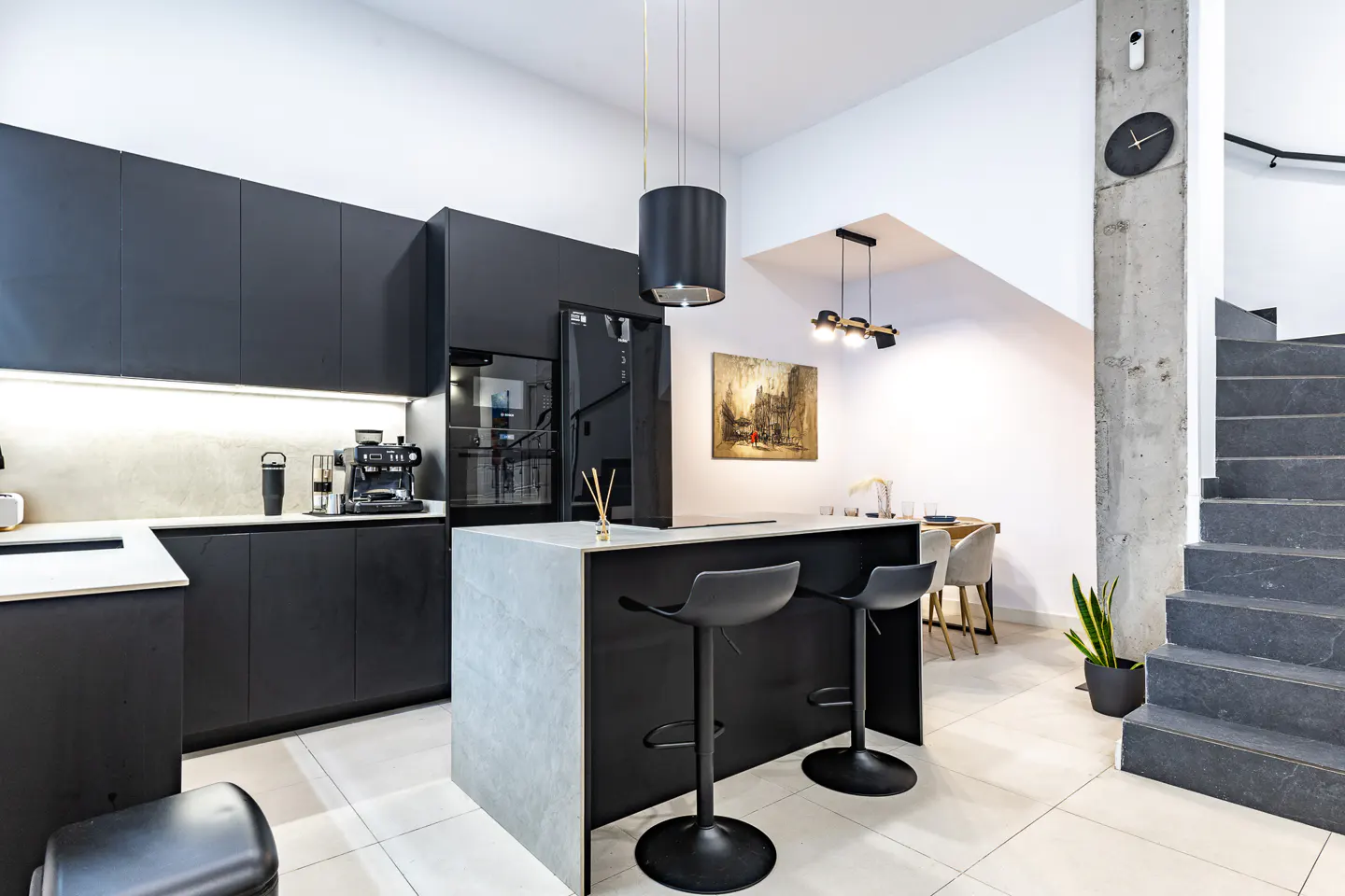 Modern kitchen with matte black cabinets, a grey island with two stools, and a staircase against a concrete pillar.