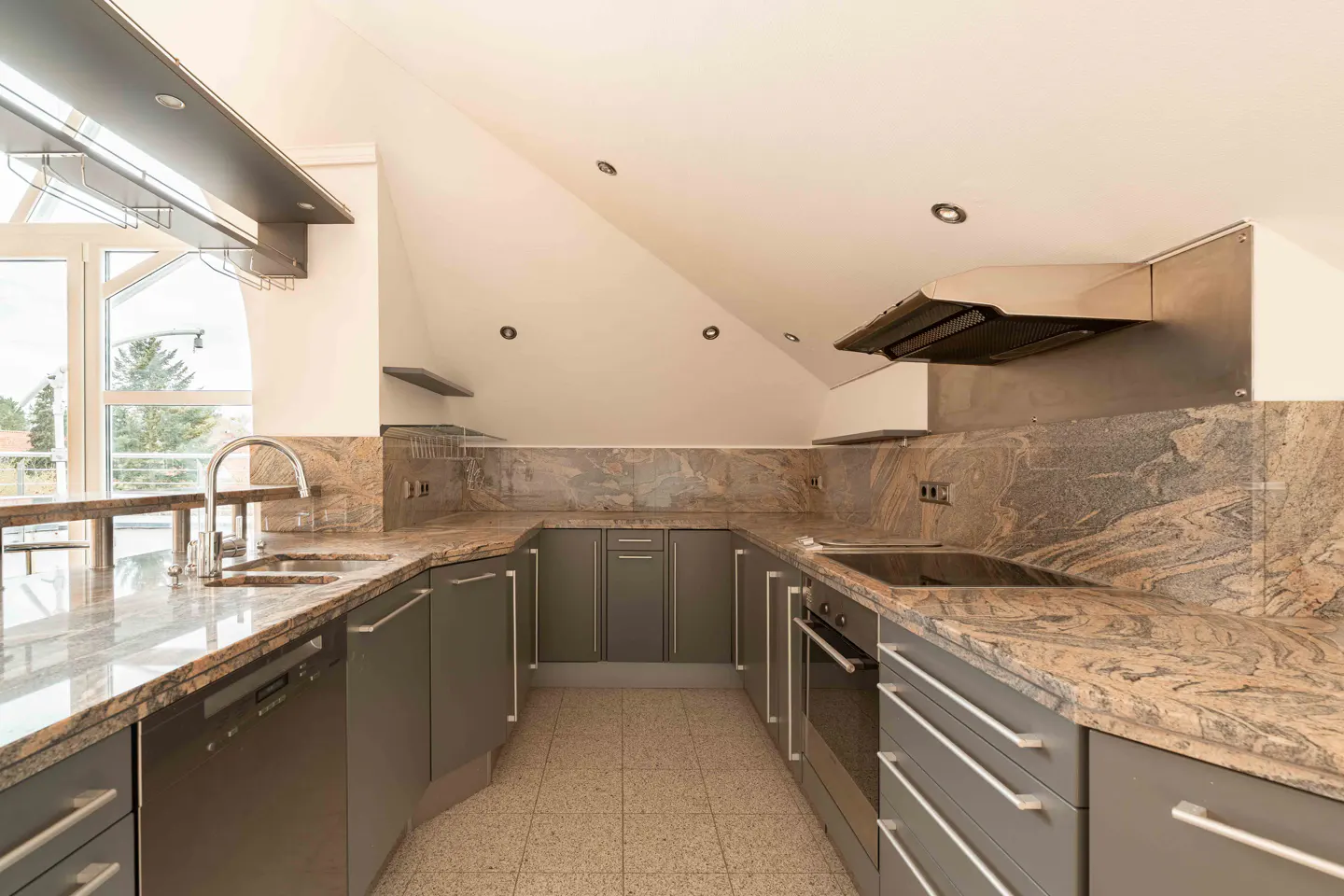 A modern kitchen with gray cabinets, granite countertops, and stainless steel appliances. A large window provides natural light.