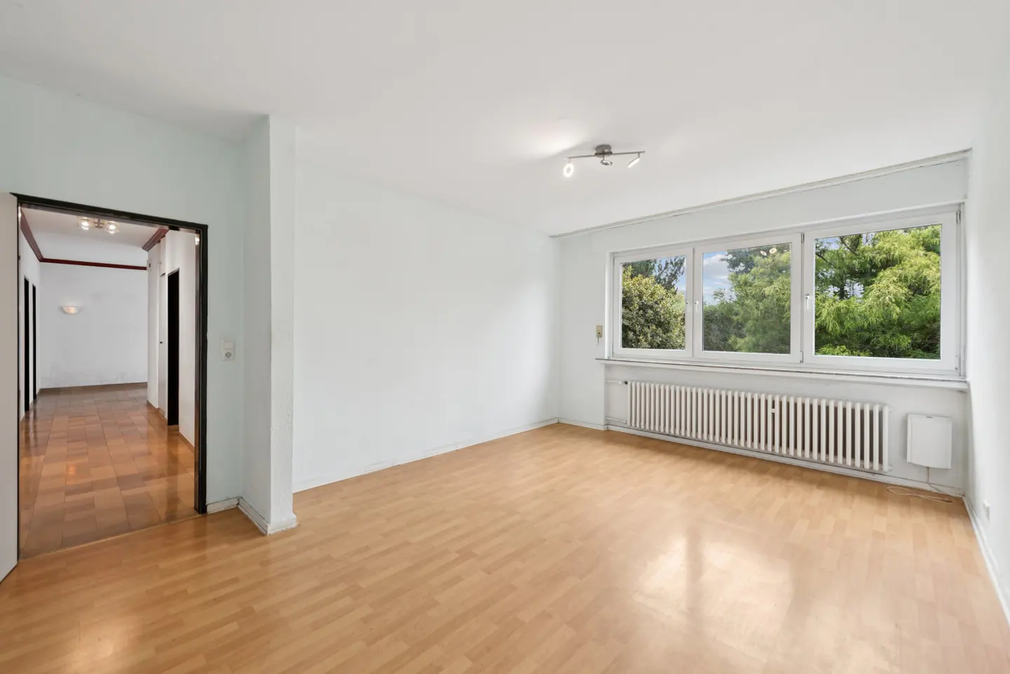 Bright, empty room with wood floors, white walls, and a large window showing green trees. A hallway is visible through a doorway.