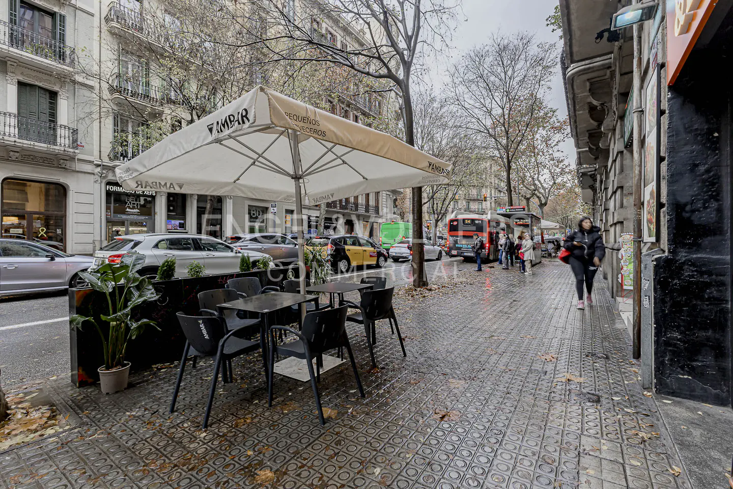 Outdoor cafe with black tables and chairs under a white umbrella on a wet city street with people and cars.