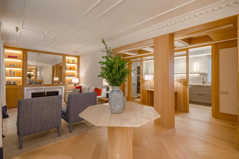 A bright living room with herringbone wood floors, gray chairs, and a marble table with a vase. Built-in shelves and a fireplace are visible.