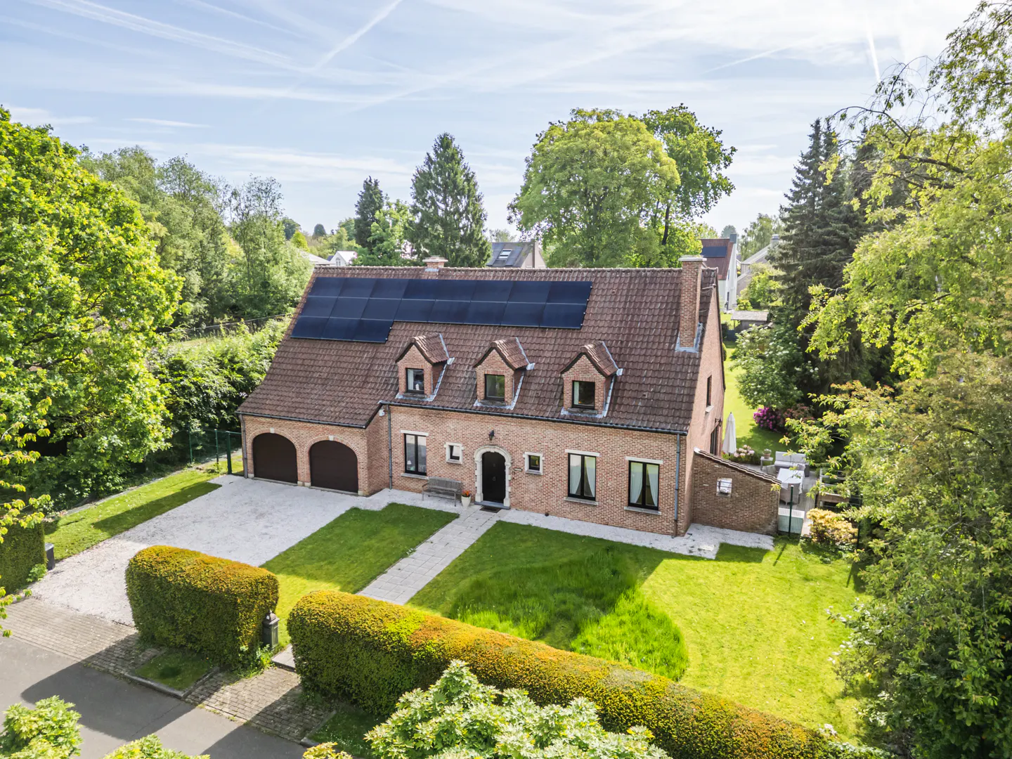 Two-story brick house with a red tile roof and solar panels, surrounded by green trees and a lawn.