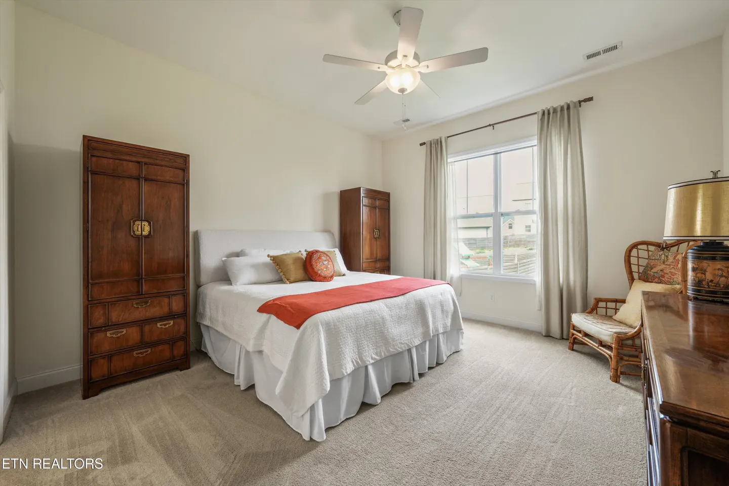 Bedroom with a white bed, orange blanket, and wooden furniture. A ceiling fan hangs above. Neutral carpet and curtains.