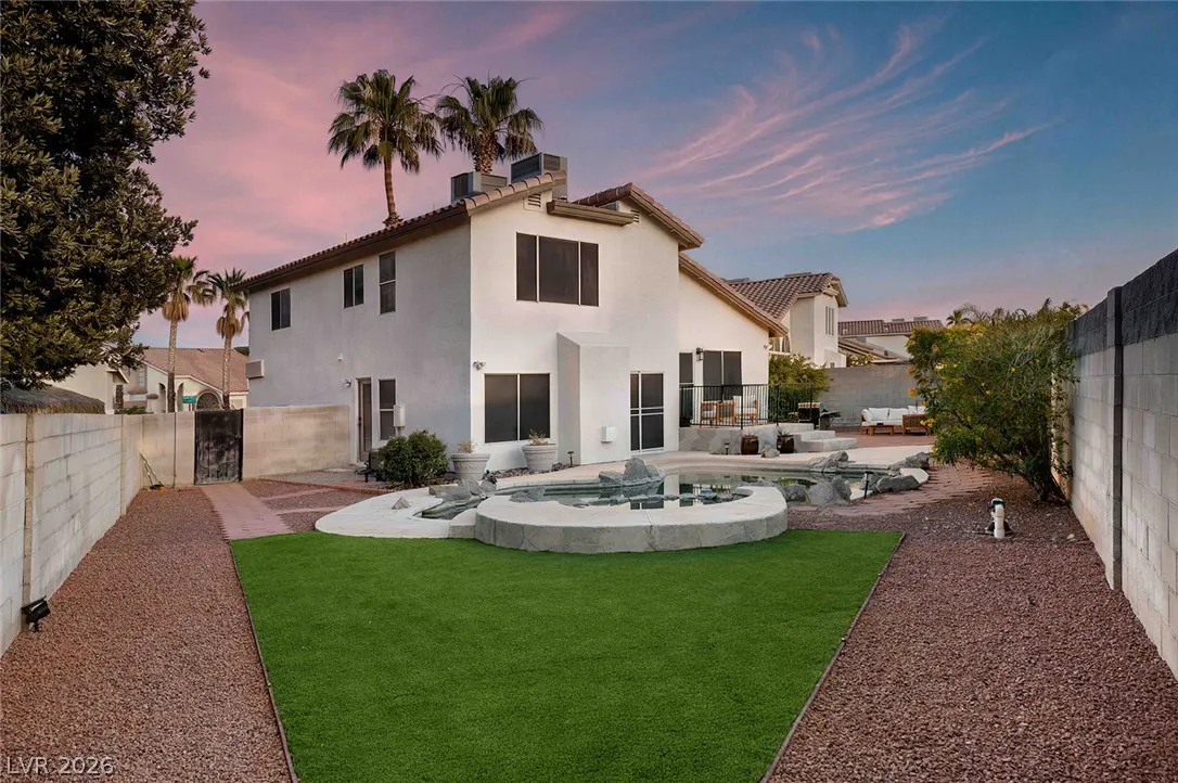 Backyard of a white two-story house at sunset, featuring a rock-lined pool, green turf, and gravel paths.