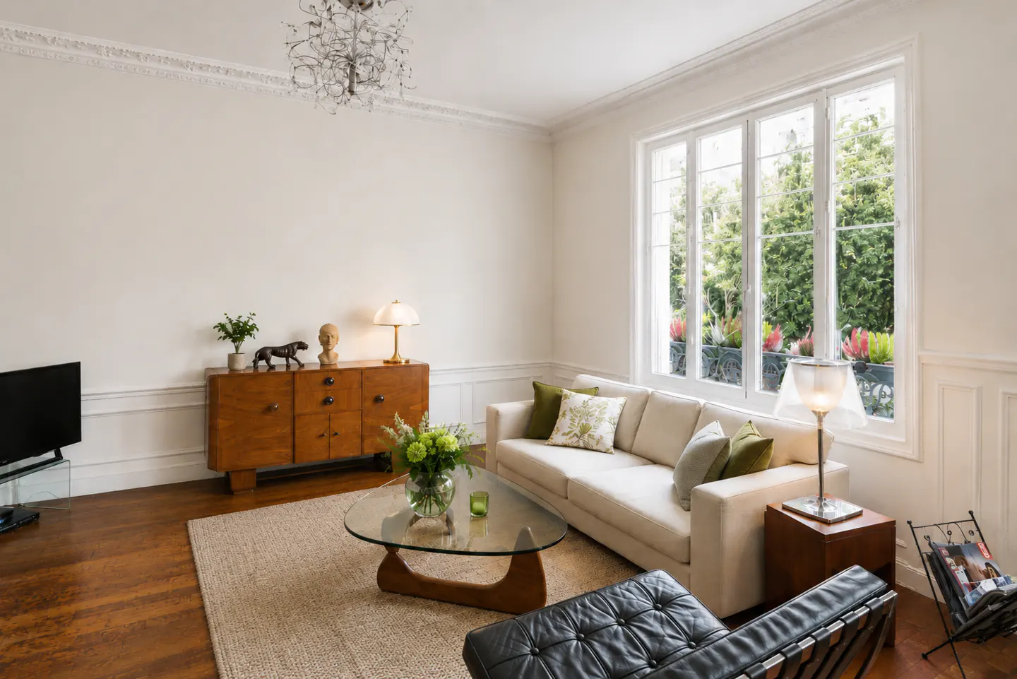 Bright living room with white walls, wood floors, and a large window overlooking greenery. A beige sofa, glass table, and wooden cabinet furnish the space.
