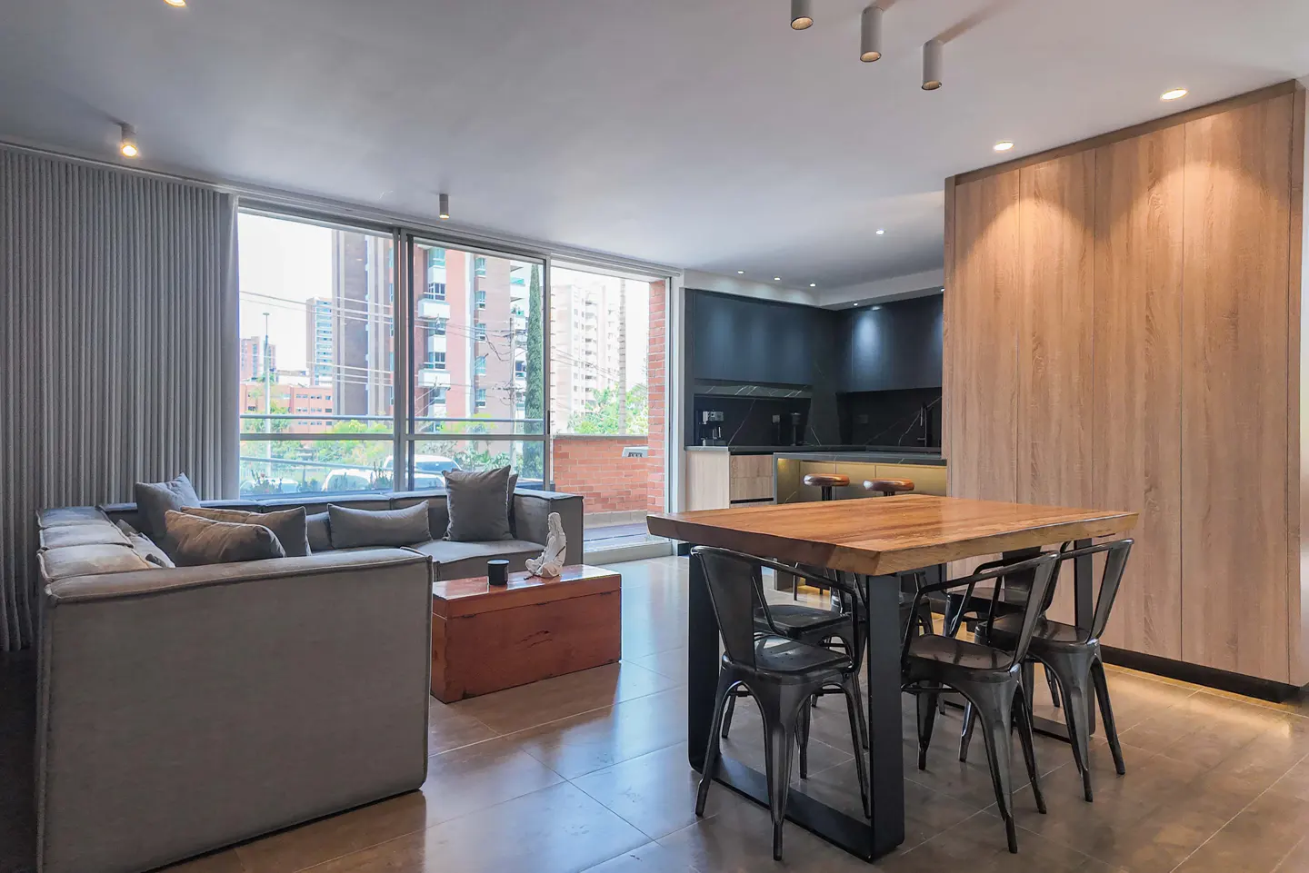 Modern open-plan apartment with a grey sectional sofa, wooden coffee table, and dining area with metal chairs.