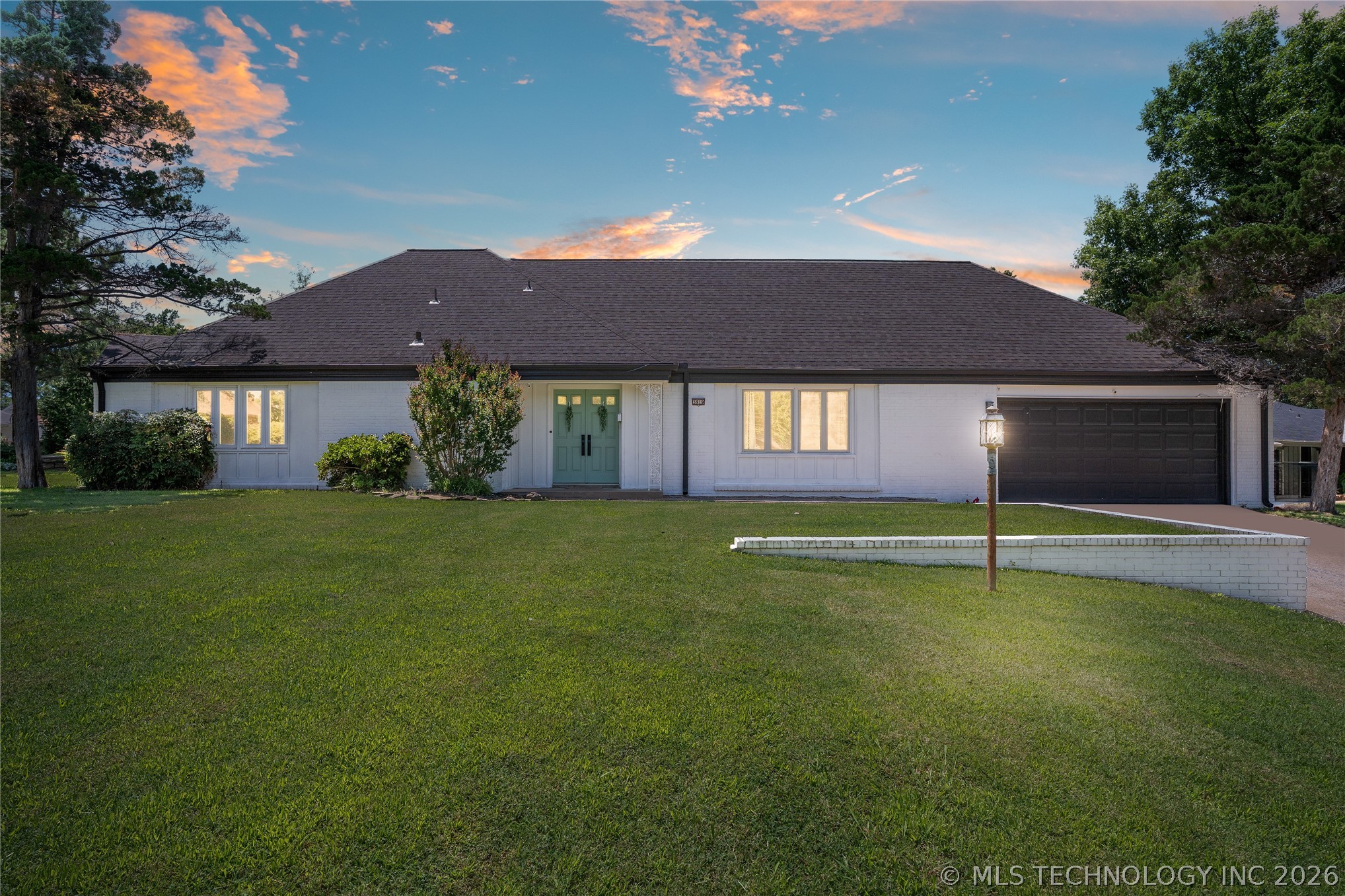 Single-story white ranch home with a dark roof, sage green front door, and large lawn under a sunset sky.