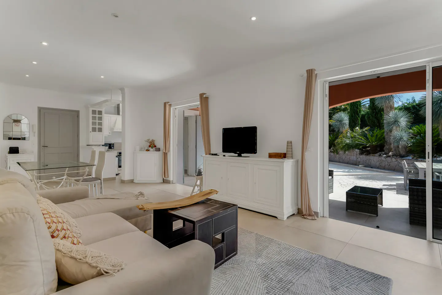 Bright living room with a beige sofa, dark wood coffee table, TV on a white cabinet, and sliding doors to a patio garden.