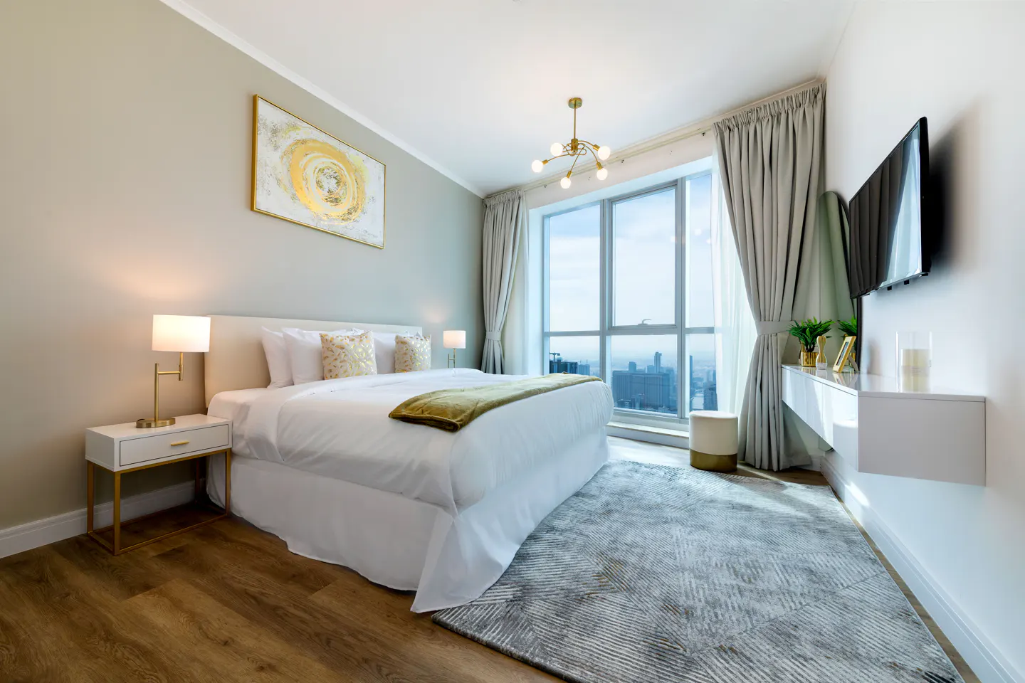 Bright bedroom with a white bed, gold accents, and a large window overlooking a city skyline. A gray rug covers the wood floor.