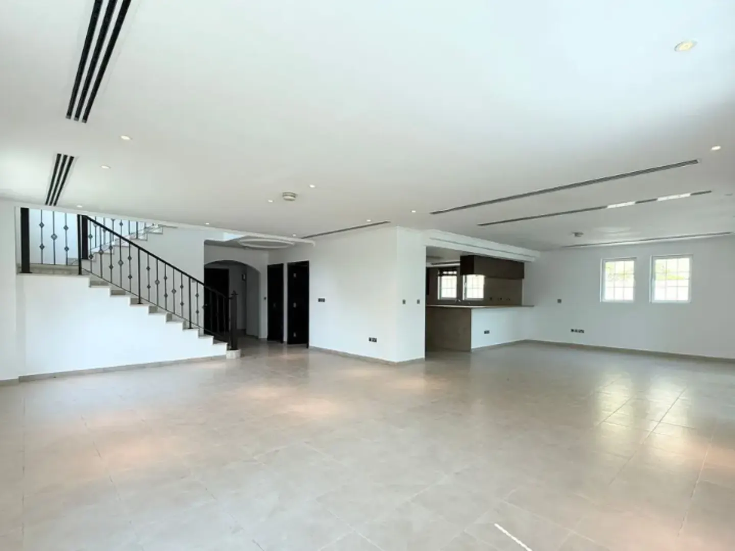 Bright, empty living room with tile floor, white walls, and staircase. Kitchen area visible in the background.