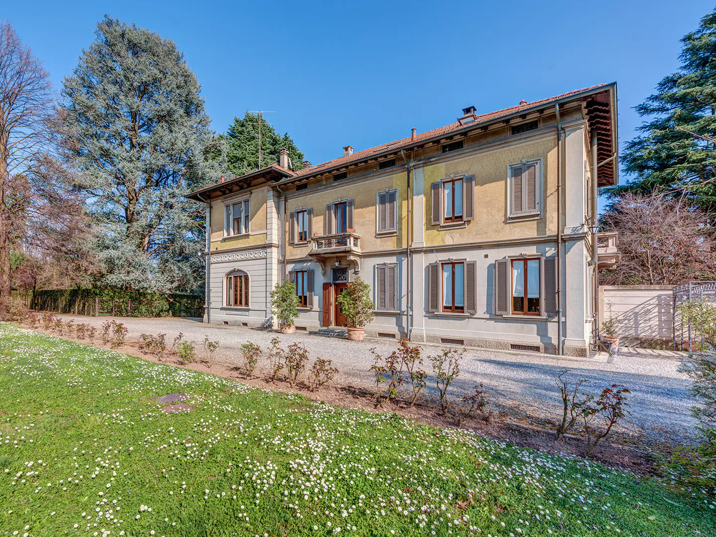 Two-story yellow house with brown shutters, a small balcony, and a green lawn with white flowers.