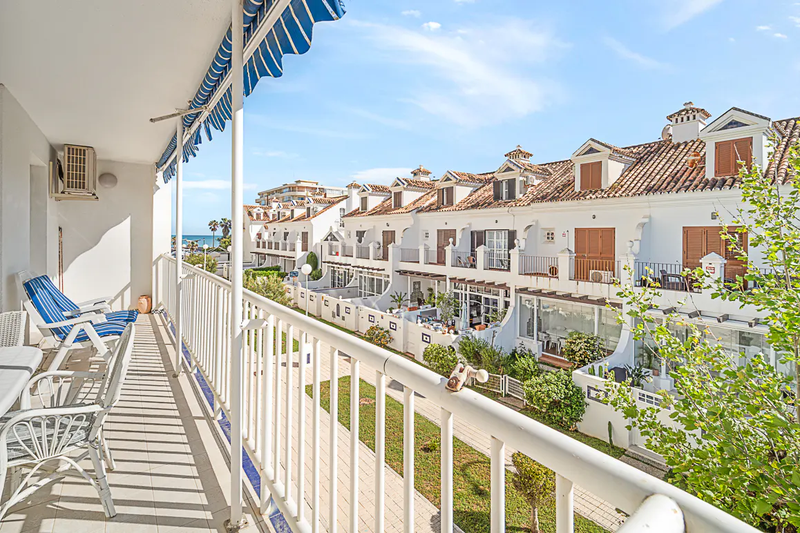 Balcony view of white townhouses with red tile roofs under a blue sky. White railings and blue striped awning. Chairs and table on the balcony.