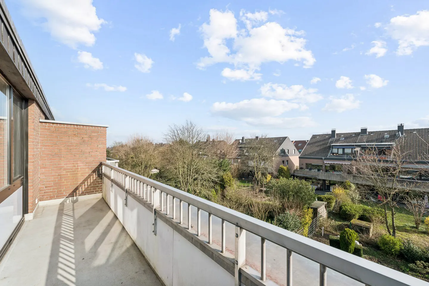 A balcony with a metal railing overlooks a neighborhood with houses, trees, and a blue sky with white clouds.