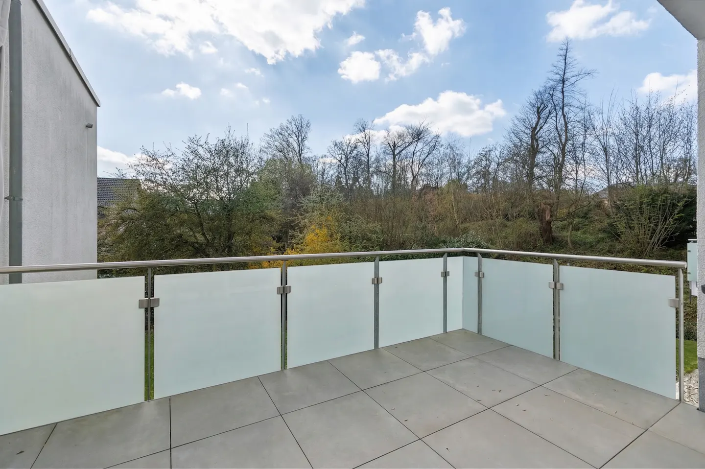 Balcony with frosted glass railing and gray tiled floor overlooks trees under a blue sky with clouds.