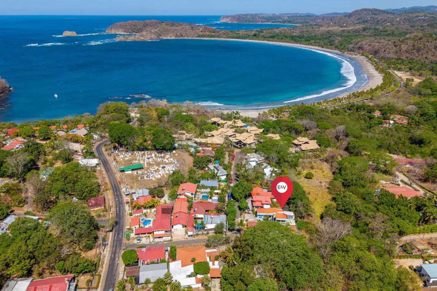 Aerial view of a coastal town with red-roofed houses, lush green trees, and a sandy beach curving around a blue bay. A red location pin marks a property.