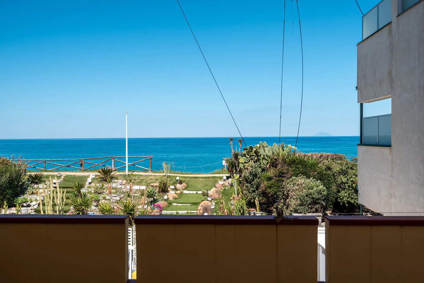 View from a balcony overlooking a garden, the ocean, and a clear blue sky. A white building is on the right.