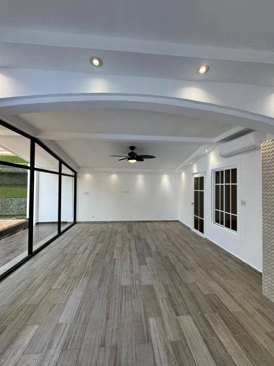 Bright, empty room with wood-look tile floor, white walls, black-framed windows, and a ceiling fan.