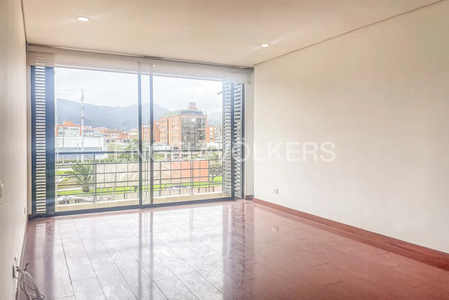 Empty room with wood floors and white walls. Large window with city view and balcony.