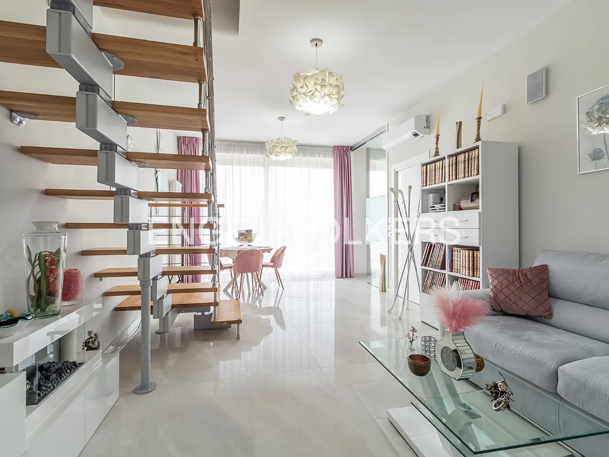 Bright, modern living room with a spiral staircase, dining table, and gray sofa. White tile floors reflect light from the window.
