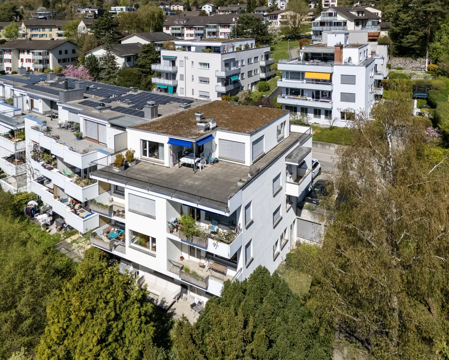 Aerial view of a modern white apartment building with balconies and a rooftop terrace, surrounded by green trees.
