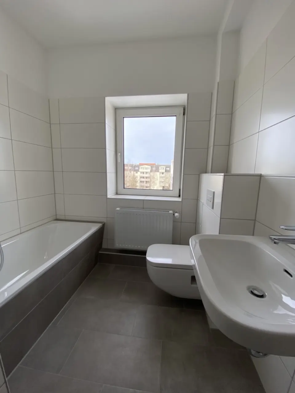 Modern bathroom with white wall tiles, gray floor tiles, a bathtub, a wall-mounted toilet, and a sink under a window.