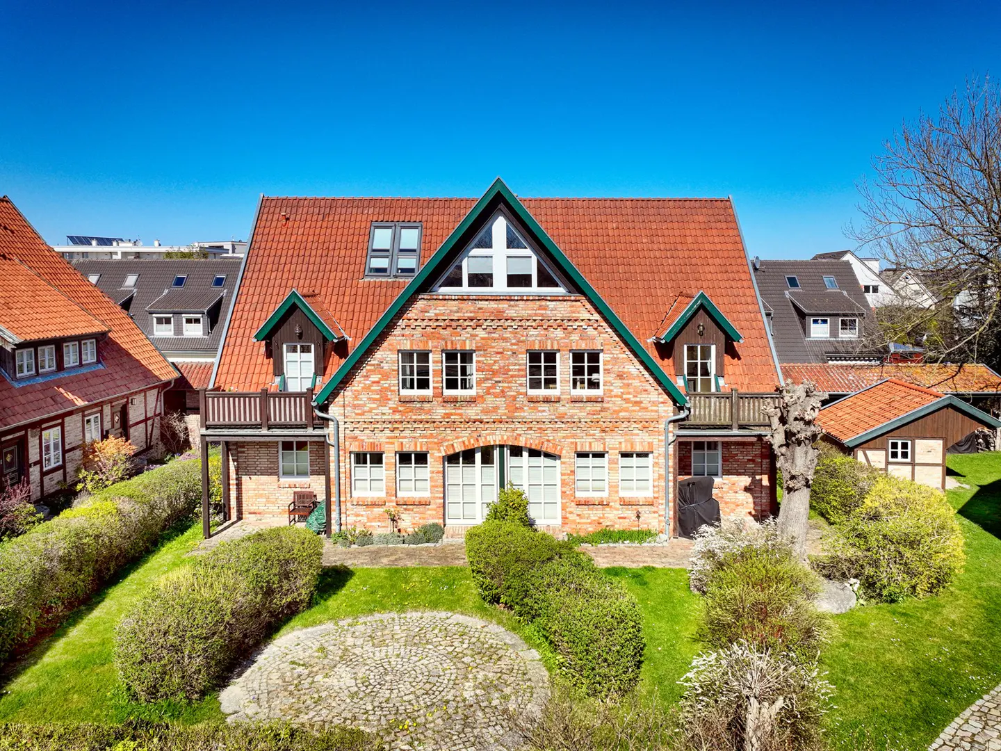 Two-story brick house with a red tile roof and green trim, surrounded by a green lawn and bushes under a clear blue sky.