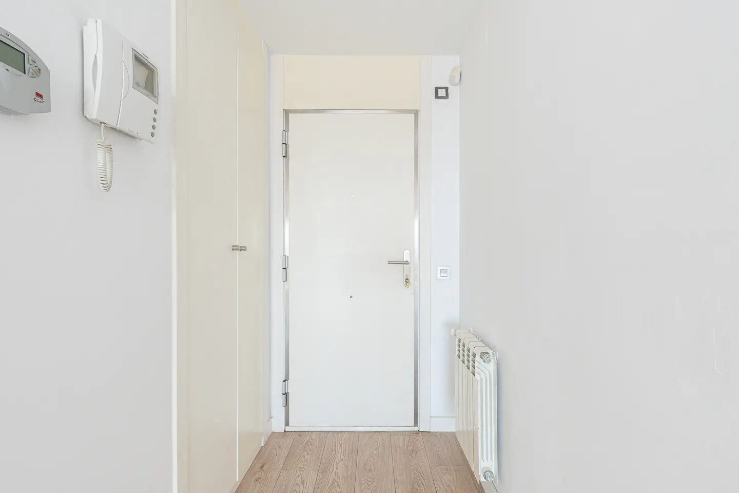 A bright hallway with white walls, light wood flooring, a white door, a wall-mounted intercom, and a white radiator.