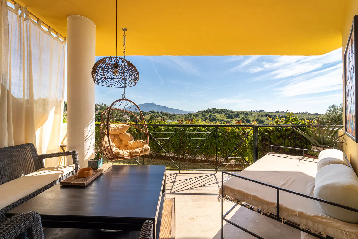 Outdoor patio with yellow ceiling, hanging chair, table, and daybed overlooking green hills and blue sky.