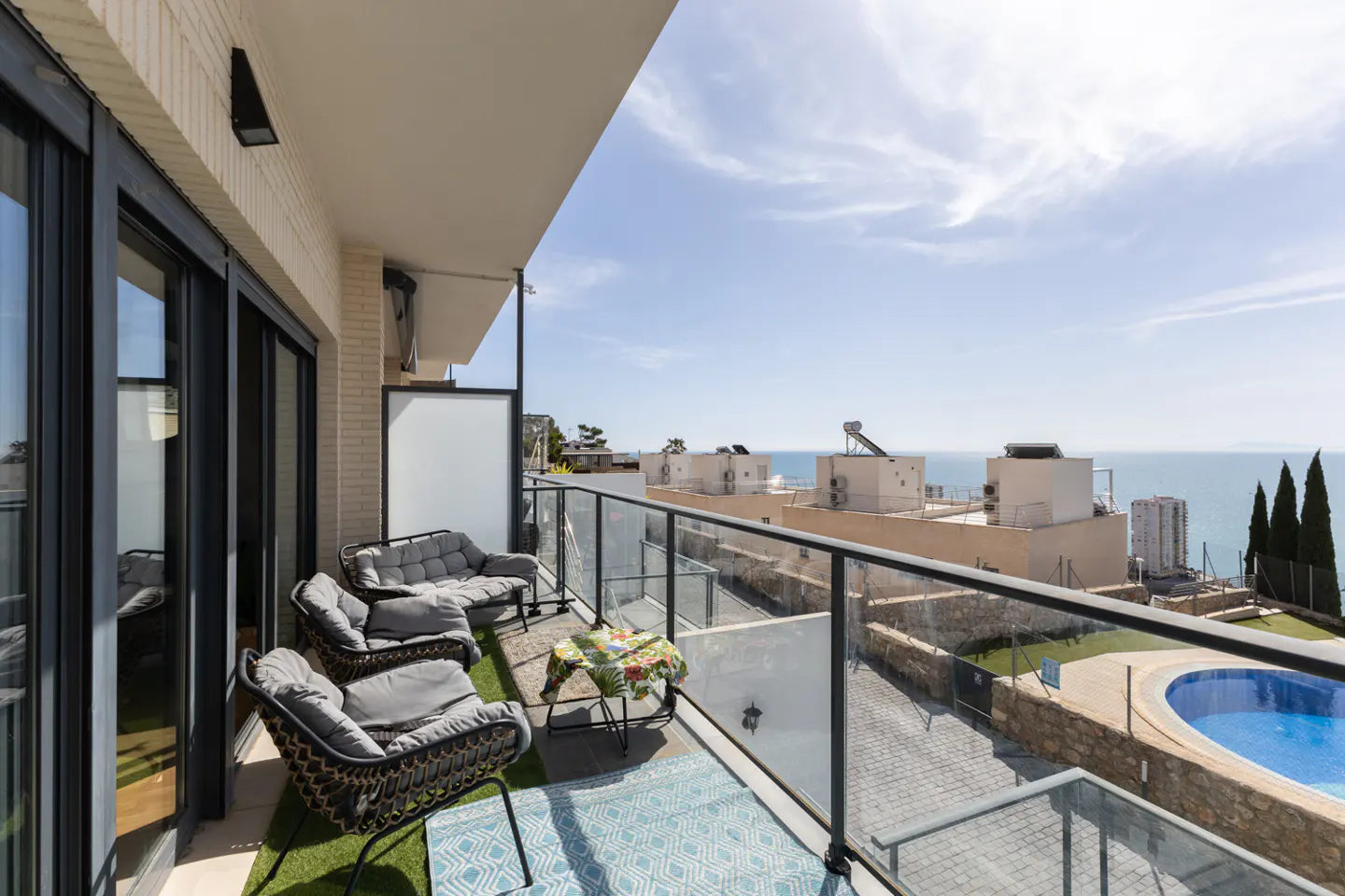 Balcony with gray wicker furniture, blue rug, and glass railings overlooking a pool, buildings, and the ocean under a blue sky.