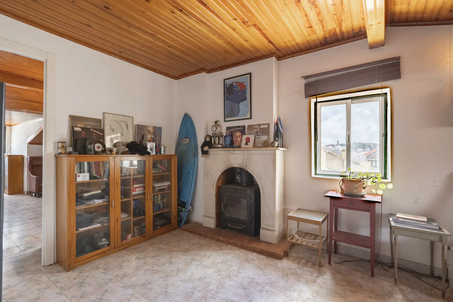 Cozy living room with wood-paneled ceiling, tiled floor, and white walls. A blue surfboard leans against a wooden cabinet next to a fireplace.