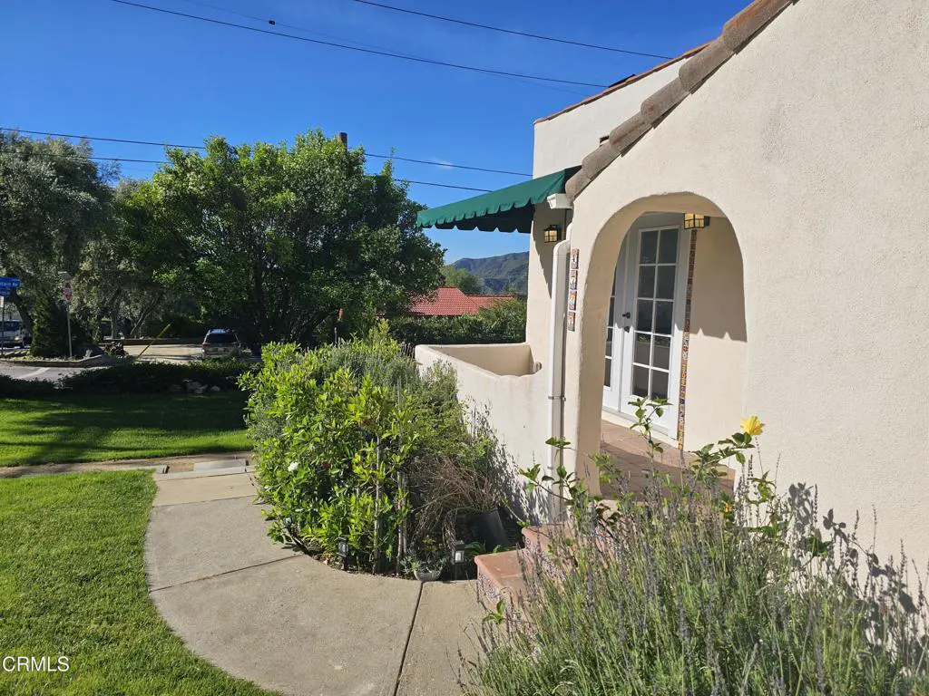 Exterior view of a stucco house with a green awning, arched doorway, and tile roof. Lush greenery and a sidewalk lead to the entrance.