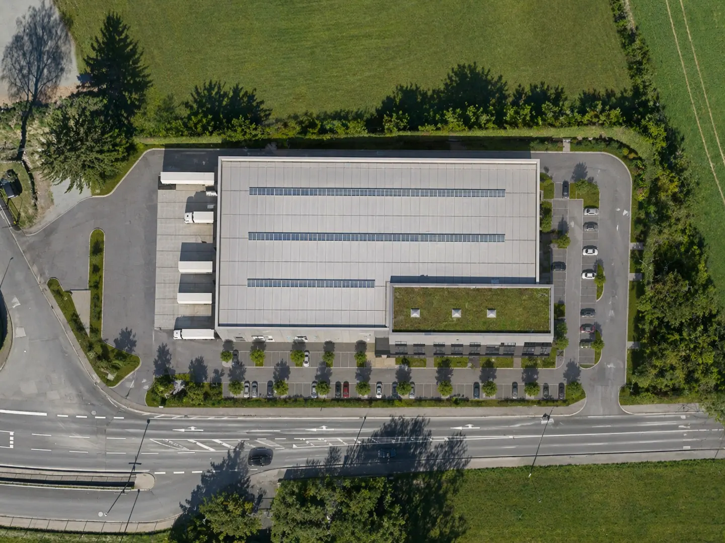 Aerial view of a gray warehouse with a green roof, loading docks, parking, and surrounding green fields.