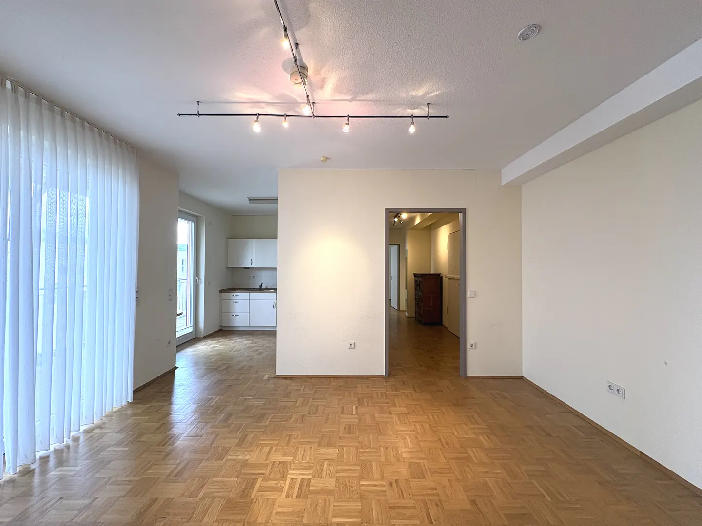 Bright, empty apartment with parquet floors, white walls, and track lighting. A kitchen and hallway are visible through openings.