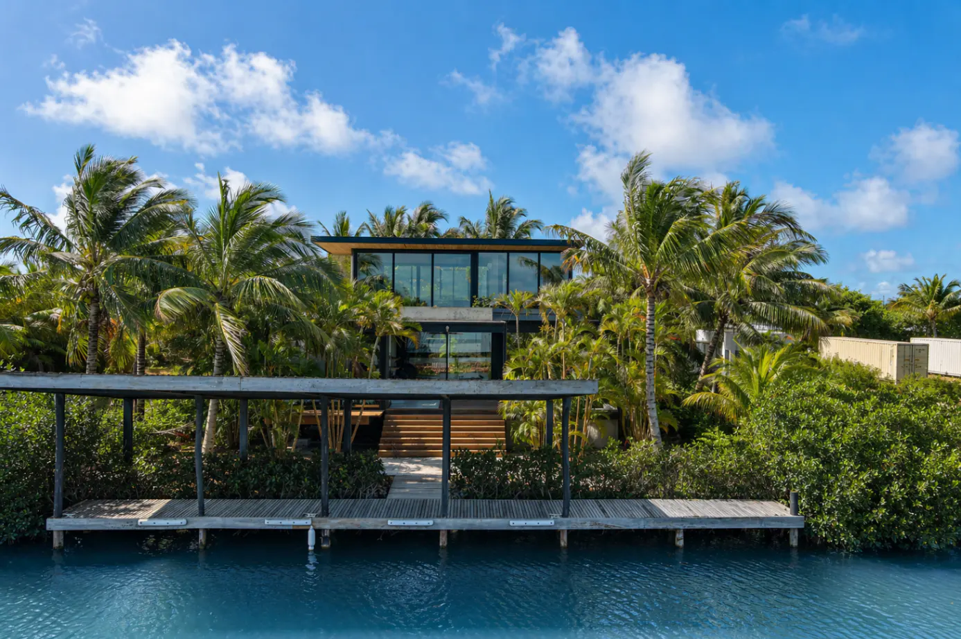 Modern waterfront home with a dock, surrounded by lush tropical greenery and palm trees under a blue sky with scattered clouds.
