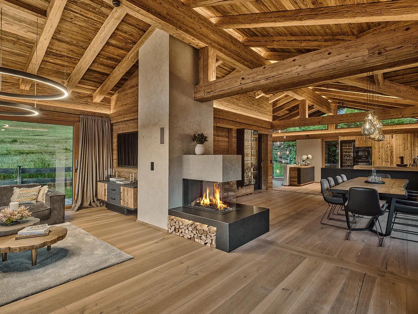 Modern open-plan living area with wood-beamed ceilings, a central concrete fireplace, dining table, and mountain views.