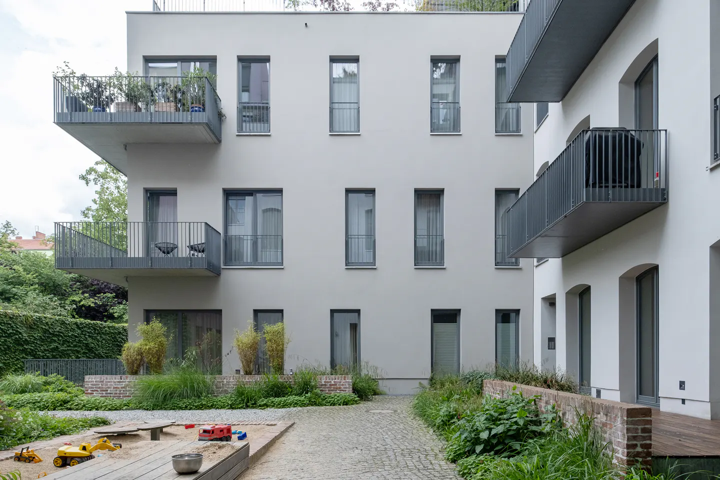 Modern apartment building with gray balconies overlooking a courtyard with a sandbox and greenery.