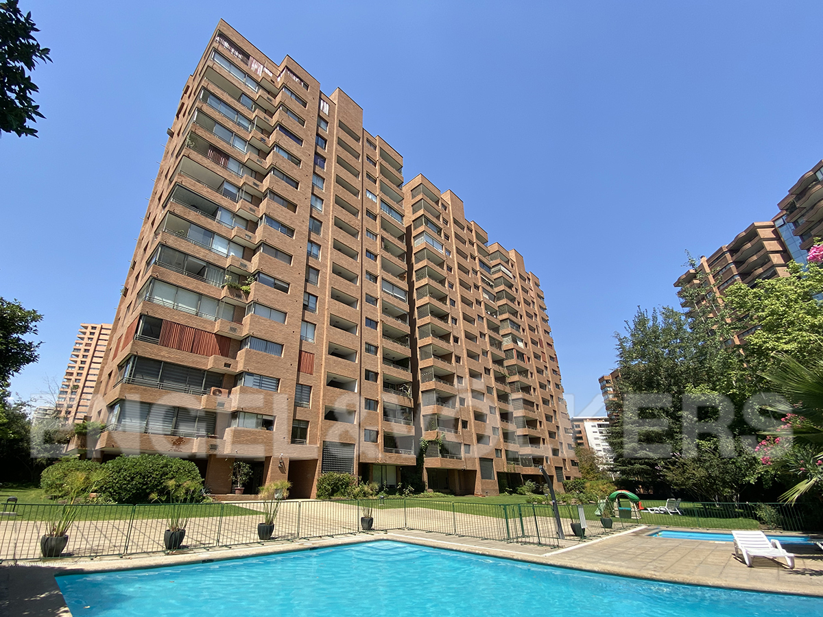 Tall brown brick apartment building with balconies overlooking a blue swimming pool and green lawn under a clear blue sky.