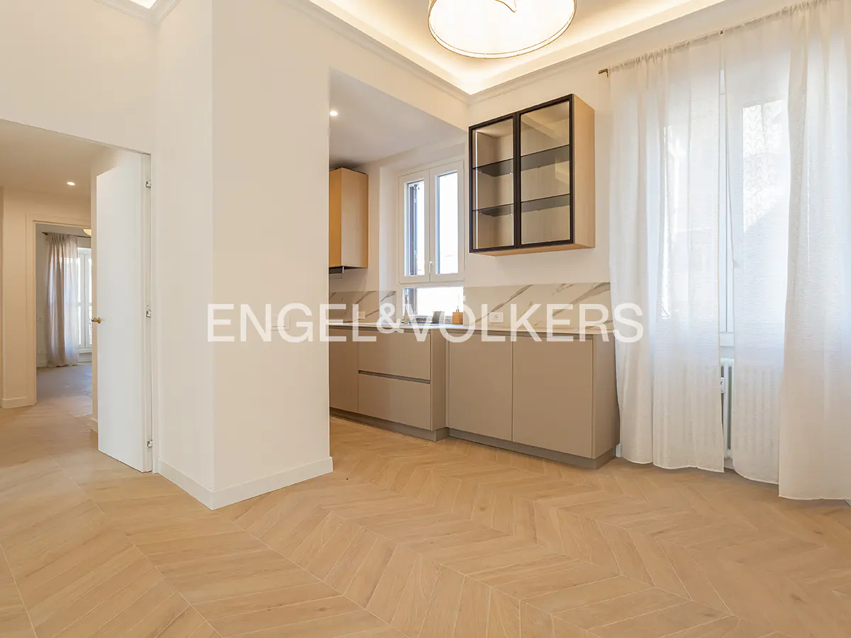 A bright, modern kitchen with beige cabinets, marble countertops, and herringbone wood floors. White sheer curtains filter light from a window.