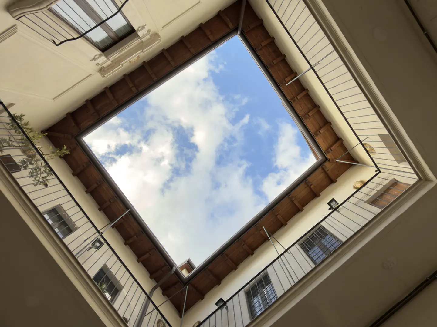 Looking up at a square courtyard opening to a blue sky with white clouds. The building has white walls, windows, and black railings.