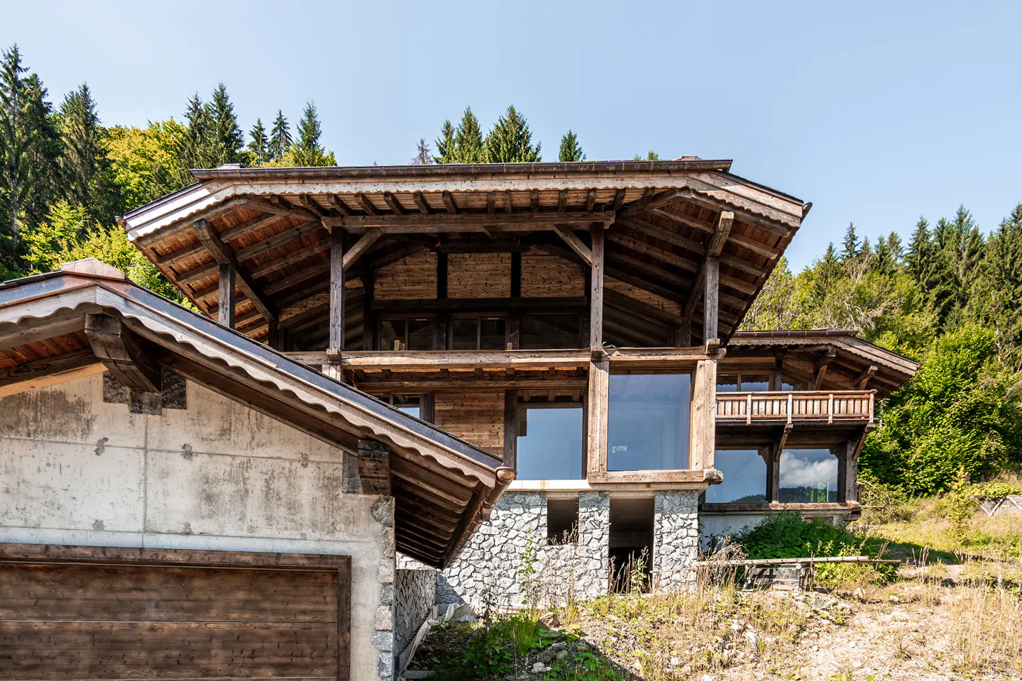 Exterior of a rustic wooden chalet with large windows, stone foundation, and forest backdrop.