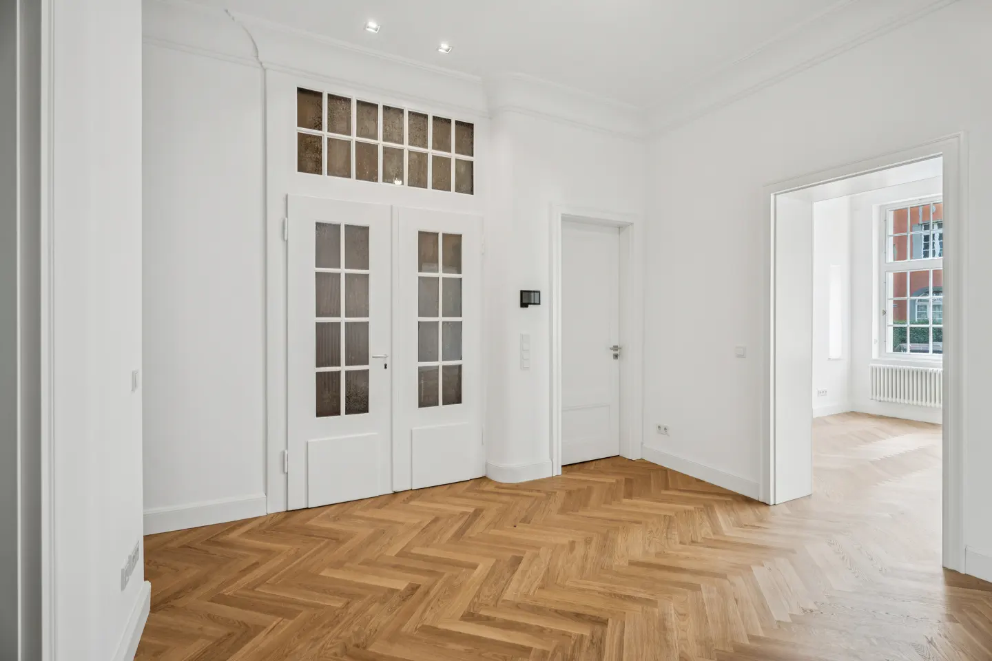 Bright room with white walls, herringbone wood floors, double glass-paneled doors, and a view into an adjacent room.