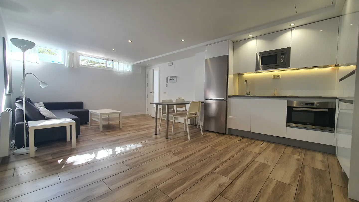 Modern open-plan apartment with wood-look floors, a grey sofa, dining area, and a sleek white kitchen with stainless steel.