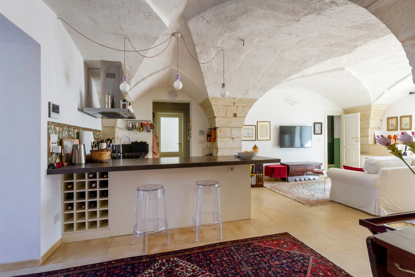 Open-concept kitchen and living room with vaulted ceilings, stone archways, and modern furnishings. Wine rack and clear stools at the kitchen island.